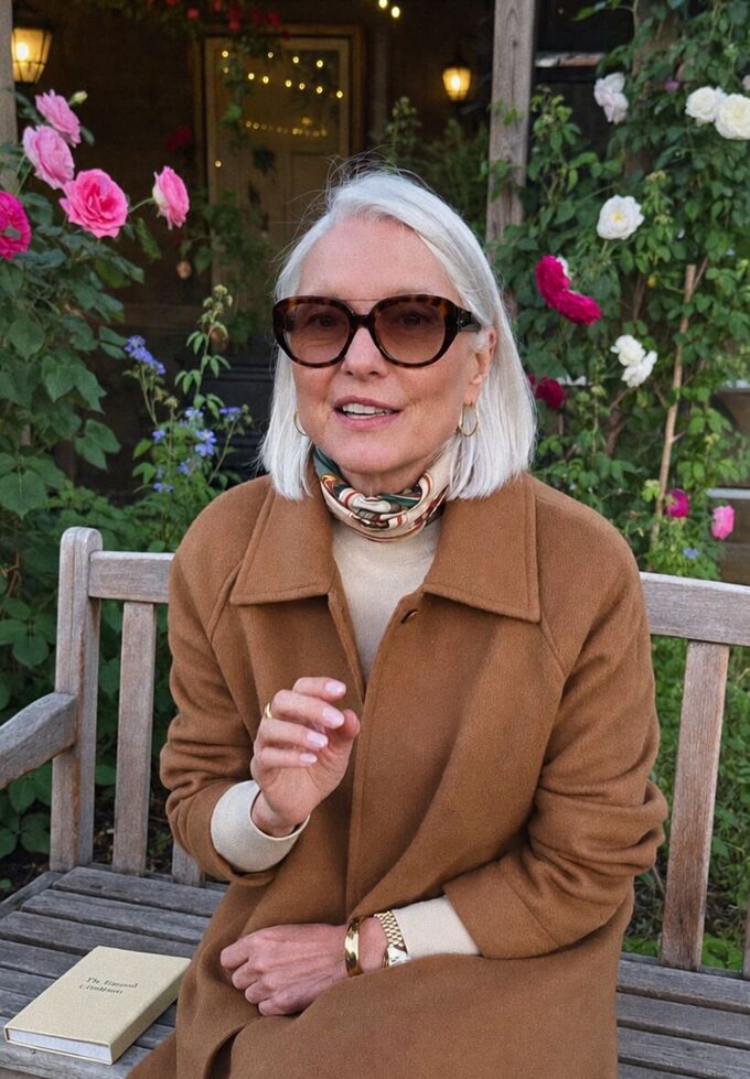 Eleanor in a camel coat and silk scarf on a garden bench among roses